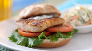 A grilled salmon burger with lettuce and tomato on a whole grain bun, served with a side of coleslaw and a beer in the background.
