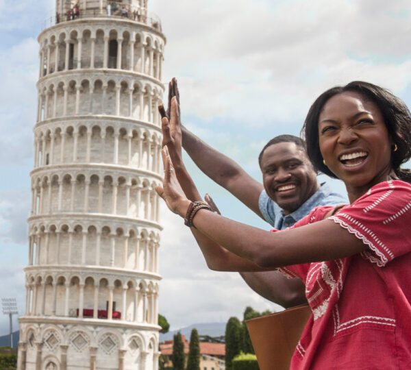 IMG-6_Europe A man and woman pose playfully in front of the Leaning Tower of Pisa, pretending to hold it up with their hands, smiling joyfully.