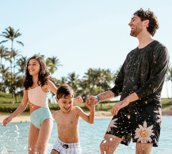 IMG-8_Mexico A man and two children play in shallow water, laughing and splashing, surrounded by palm trees and a sunny sky.