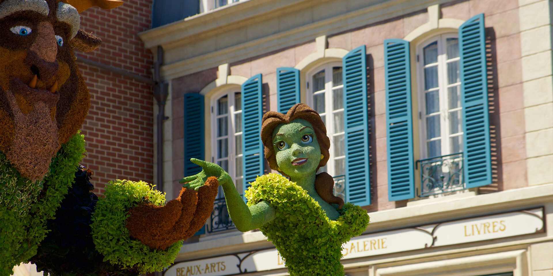 A large, whimsical sculpture of a beast and a princess made of greenery, in front of a building with blue shutters.