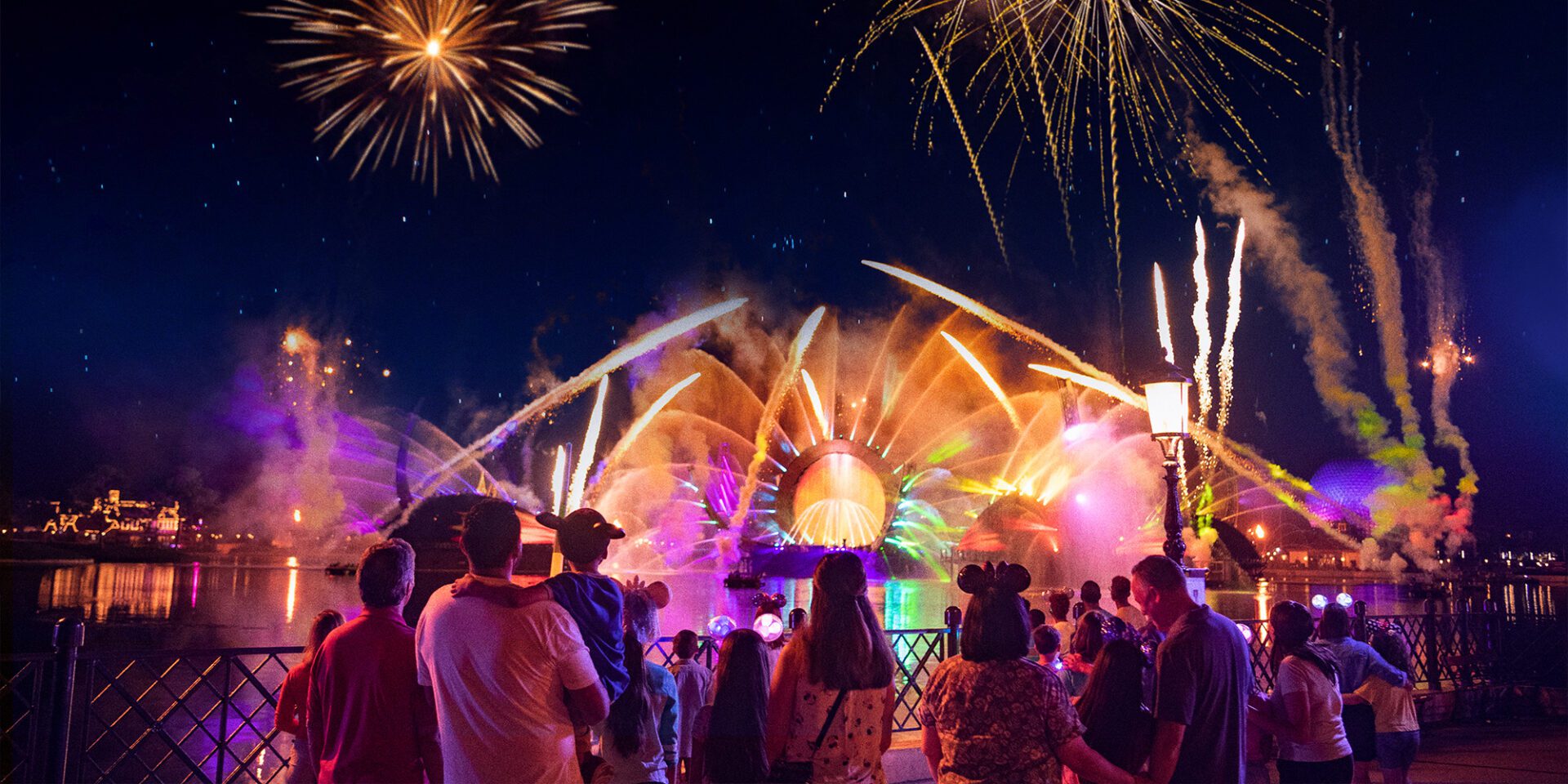 A vibrant nighttime scene with colorful fireworks above a crowd by a water feature, illuminated in hues of purple and blue.
