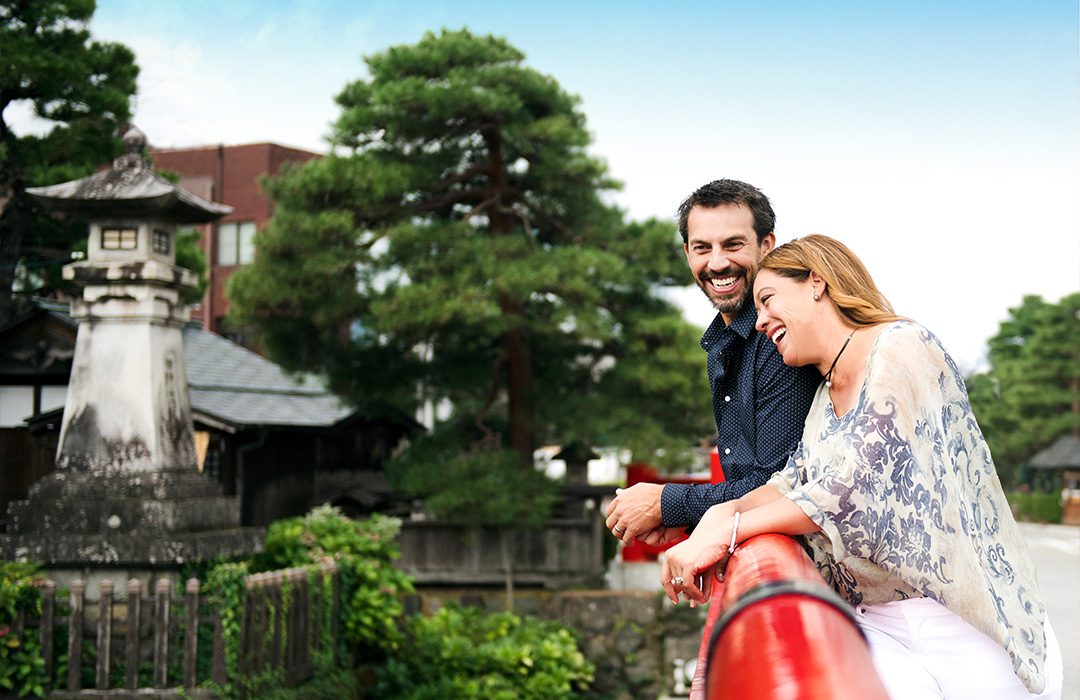 content 2-abd A smiling couple leans over a red bridge, surrounded by greenery and traditional architecture, enjoying a scenic view.