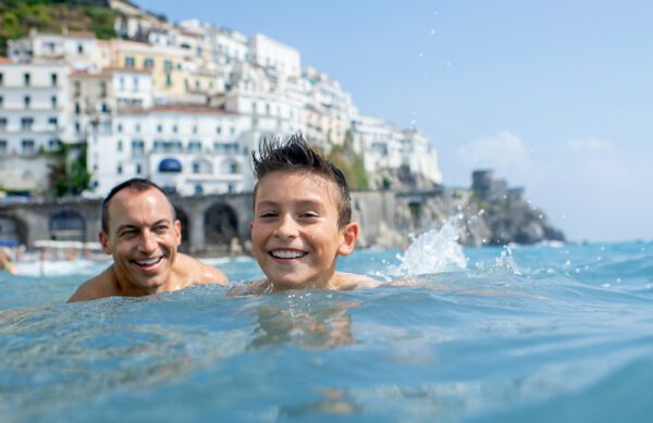 content 4-abd A man and a boy swim in clear water, smiling, with white buildings and a cliff in the background under a bright blue sky.