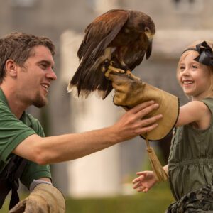 A smiling man in green attire interacts with a young girl, who joyfully holds a hawk on a glove, both enjoying the moment outdoors.