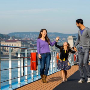 A family walks happily along a riverside promenade, enjoying the sunny day, with the cityscape in the background.