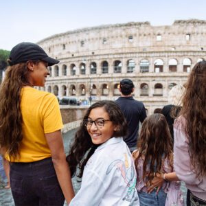 A group of people, including a smiling girl and her companion, admire the Colosseum in Rome. The scene captures a joyful family moment.