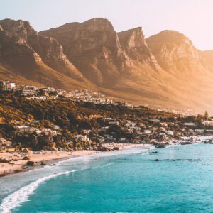 A serene beach scene with turquoise waters and sunlit mountains in the background. Coastal homes are nestled against the hillside.