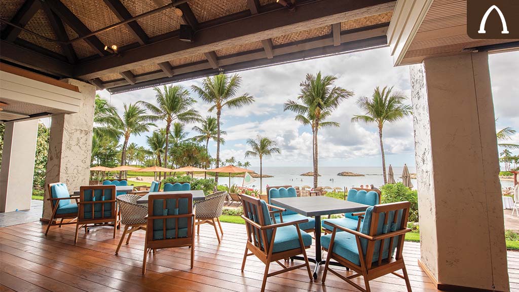 Aulani AMA AMA A tropical patio with wooden chairs and a table overlooks a beach with palm trees and cloudy skies in the background.