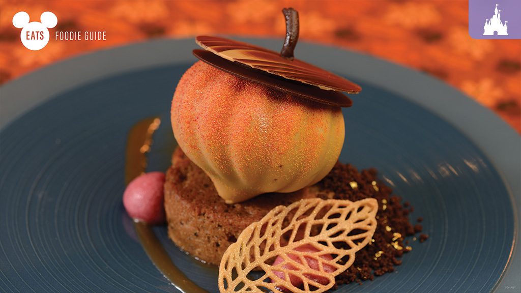 A beautifully crafted dessert resembling an apple, topped with chocolate, sits on a blue plate with decorative elements and a leaf-shaped garnish.