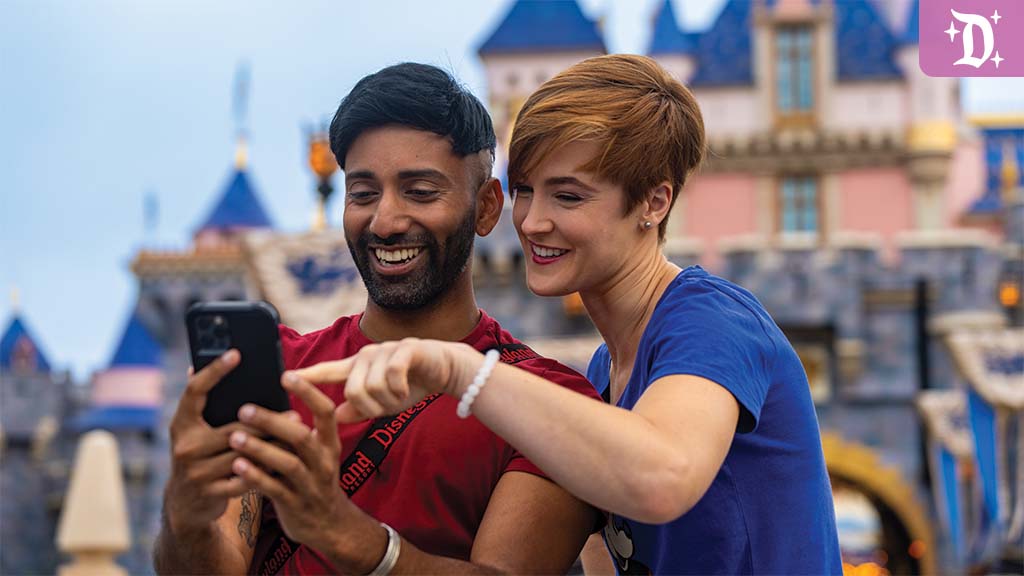 A man and woman smile and point at a smartphone, with a colorful castle in the background, enjoying their time at a theme park.
