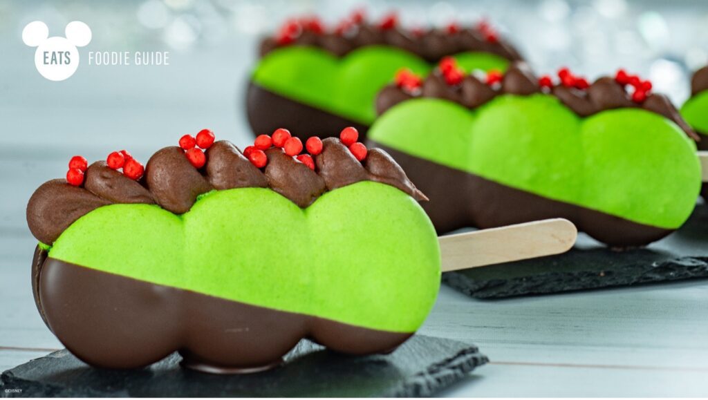 Three colorful dessert popsicles with green and brown layers, topped with chocolate frosting and red sprinkles, placed on a slate surface.