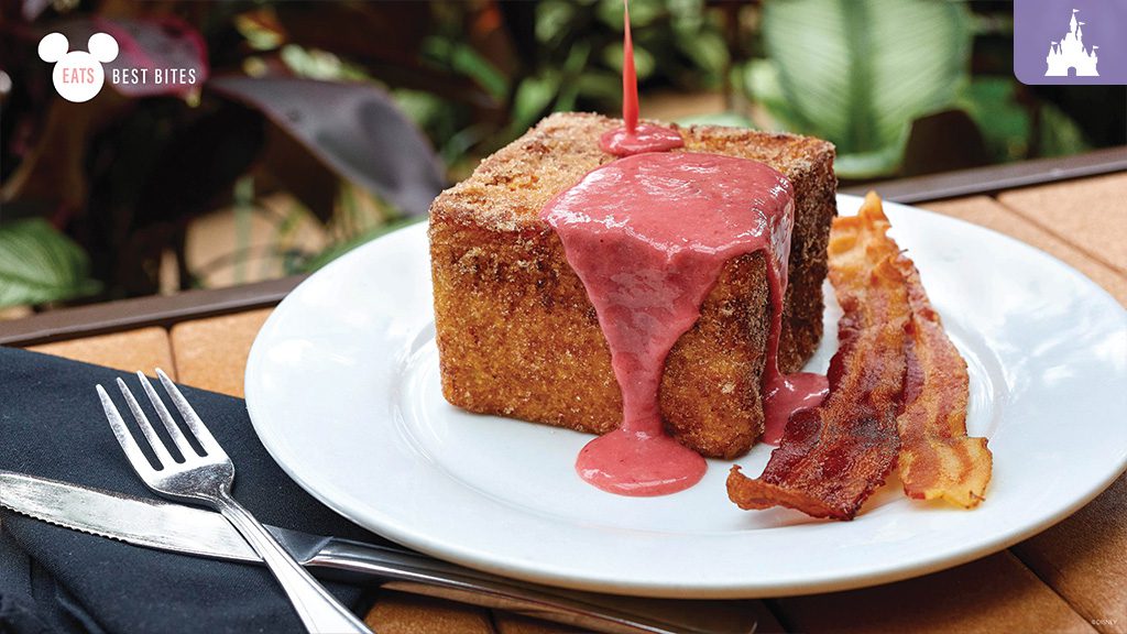 A slice of French toast topped with pink sauce and a cherry, served on a plate with crispy bacon beside it.