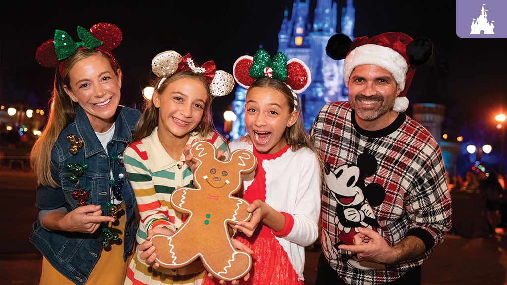 A festive group of four, wearing holiday attire and Mickey Mouse ears, smiles and poses with a large gingerbread cookie in front of a lit-up castle.