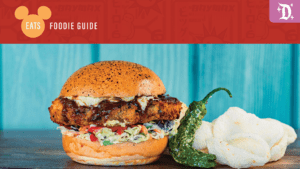 A delicious chicken sandwich topped with slaw, served with a green pepper and a side of fluffy white bread, set against a colorful background.