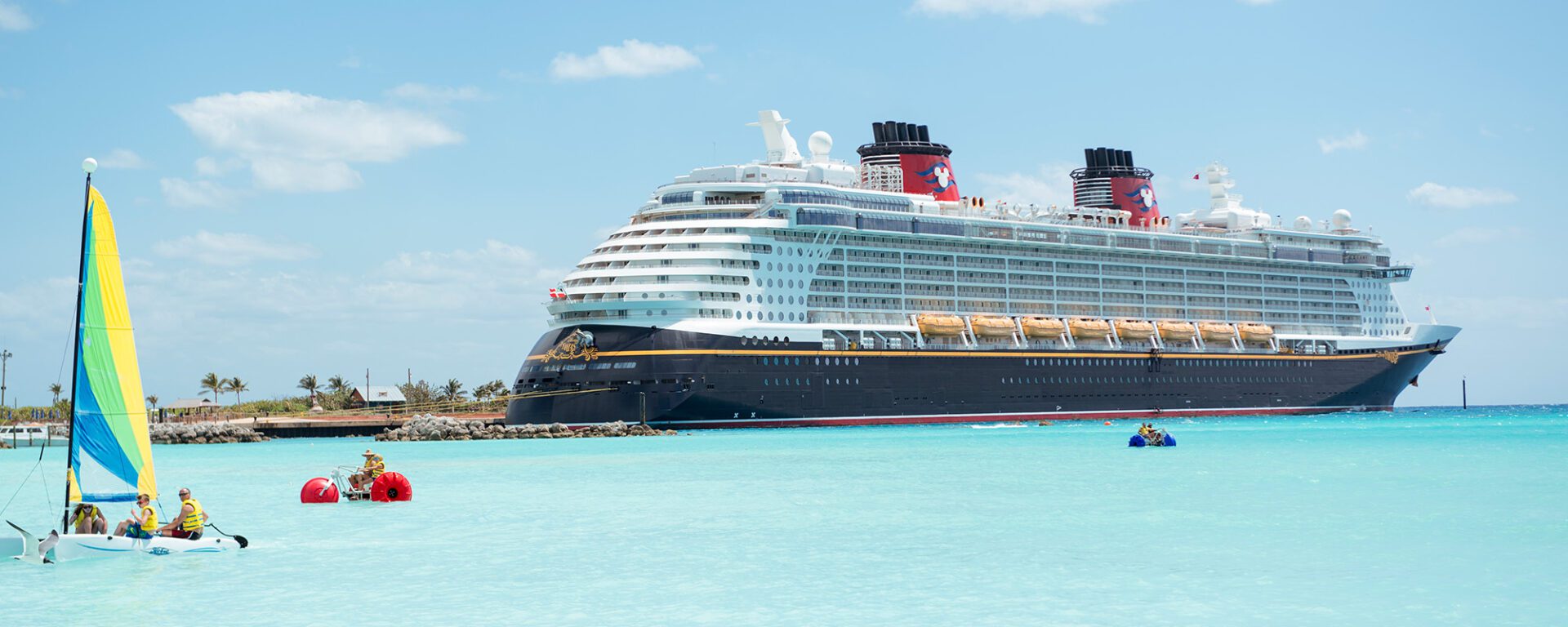 A large cruise ship docked near a sandy beach, with sailboats and kayaks on clear turquoise waters under a bright blue sky.