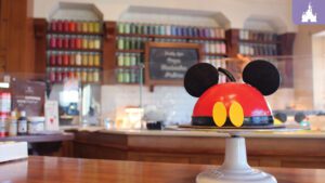 A red and black dessert shaped like Mickey Mouse's head sits on a white cake stand in a cafe with colorful shelves in the background.