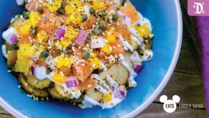 A blue bowl filled with sliced potatoes topped with salmon, mango, red onion, capers, and drizzled with creamy sauce.