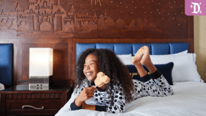 A smiling child in polka-dot pajamas lies on a hotel bed, propping herself on her elbows, with a castle mural in the background.