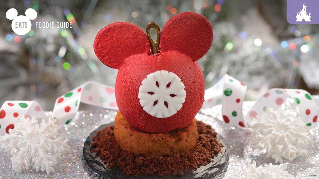 A vibrant red dessert shaped like Mickey Mouse, sitting on a crumbly base, decorated with a white circular detail and festive ribbons.