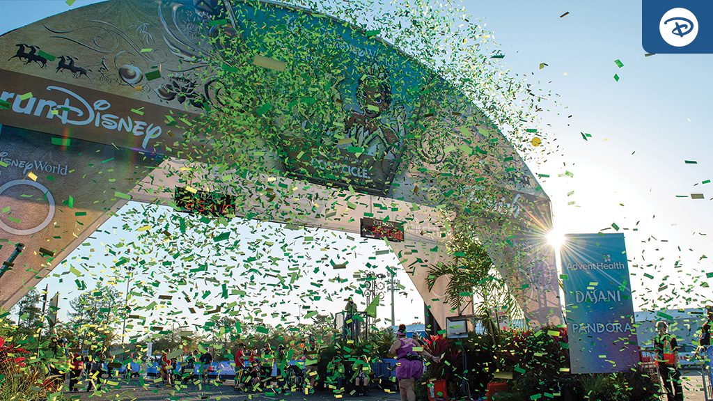 Celebratory confetti rains down at the finish line of a Disney event, with a vibrant arch and cheering crowd in the background.
