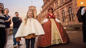 A young girl in a white cape smiles as a woman in a historical gown poses nearby, with people clapping in the background.