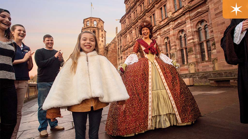 ABD 2024 River Cruise Itineraries A young girl in a white cape smiles as a woman in a historical gown poses nearby, with people clapping in the background.