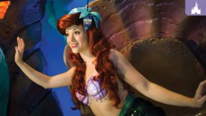 A character with long red hair, wearing a seashell top and a green tail, poses joyfully against an underwater-themed background.