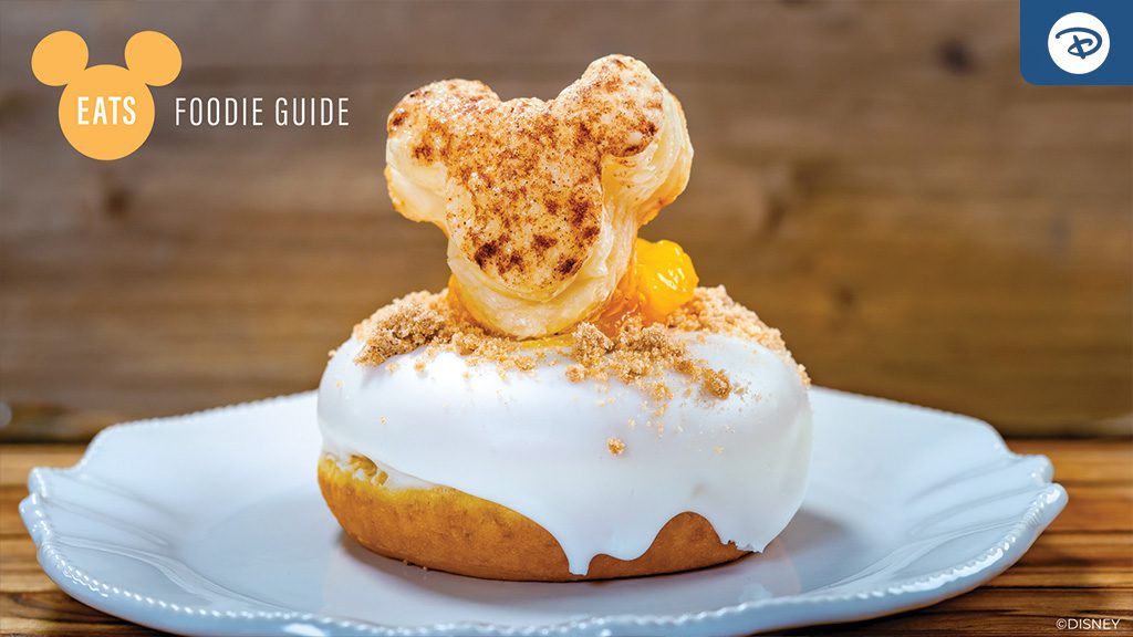 A round, frosted donut topped with a Mickey Mouse-shaped pastry, sitting on a white plate against a wooden background.