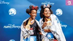 Two smiling women in Mickey Mouse ears hold medals, wrapped in silver blankets, celebrating at a runDisney event with a blue backdrop.
