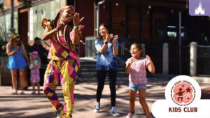 A performer in colorful attire dances joyfully alongside two children, while others look on, enjoying a festive atmosphere.