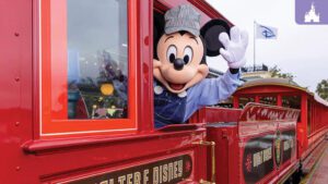 Mickey Mouse, dressed as a train conductor, waves from a red train car at a theme park, with blue skies in the background.