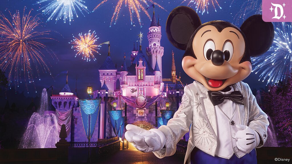 Mickey Mouse in a tuxedo smiles and poses with outstretched hand in front of a brightly lit castle and fireworks in the night sky.