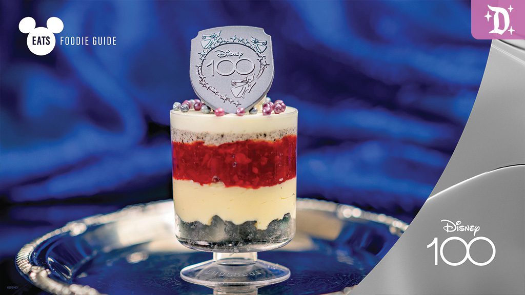 A layered dessert in a clear cup, featuring red, white, and blue layers, topped with decorative beads and a shield-shaped chocolate piece.