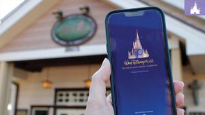 A hand holds a smartphone displaying the Walt Disney World app, with a Disney-themed building in the background.