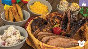 A basket filled with grilled meats, a cherry on top, surrounded by bowls of mac and cheese, potato salad, and cornbread.