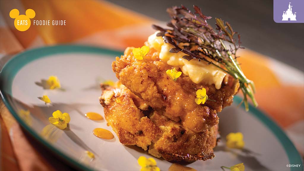 Epcot Flower and Garden Festival 2023 A gourmet dish featuring a fried stack, topped with greens and sauce, garnished with edible flowers on a green plate.