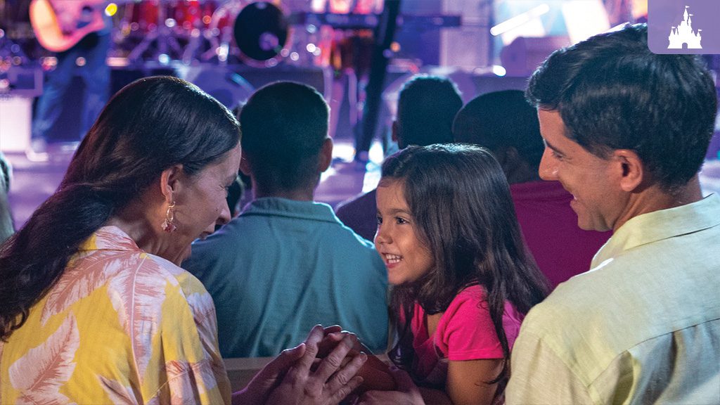 A family enjoys a live performance, smiling and interacting, with colorful stage lights illuminating the background.