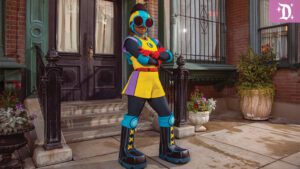 A colorful, futuristic character stands confidently on a sidewalk, wearing a vibrant outfit with oversized sunglasses, in front of a brick building.