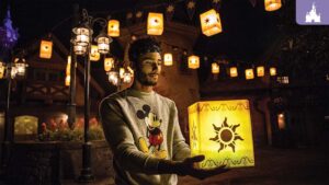 A young man holds a glowing lantern adorned with a sun design, surrounded by hanging lanterns in a nighttime setting.