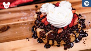 A dessert featuring chocolate cake topped with whipped cream, strawberries, and chocolate drizzle, served on a wooden platter.