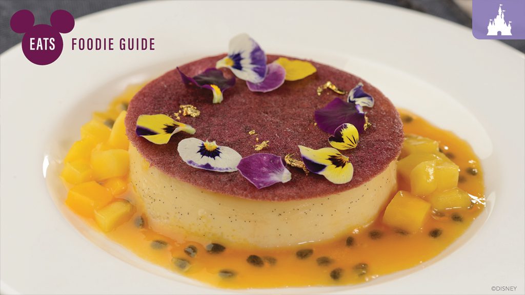 A dessert featuring a round, creamy cake garnished with edible flowers, surrounded by mango pieces and a orange sauce on a white plate.