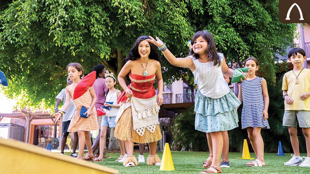 Children play outdoors, happily participating in a game, while one girl, dressed in a colorful costume, engages with them.