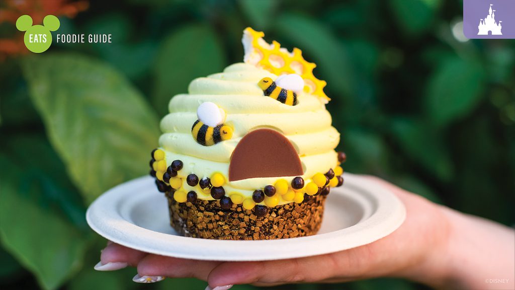 A hand holds a yellow bee-themed cupcake topped with swirls, a chocolate face, and candy decorations, set against green foliage.
