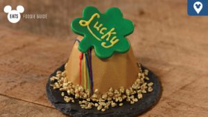 A small, rounded dessert topped with a green clover that says "Lucky," surrounded by golden sprinkles on a dark plate.