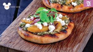 Two mini flatbreads topped with pesto, goat cheese, roasted vegetables, and fresh greens, served on a wooden board.