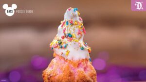A colorful dessert shaped like a Christmas tree, topped with white icing and rainbow sprinkles, set against a purple background.