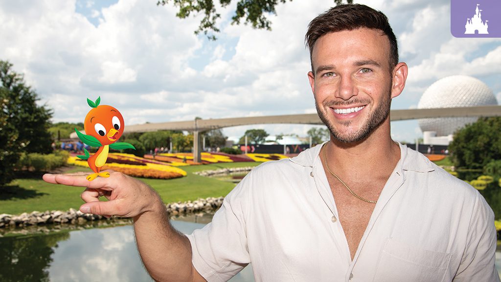 A smiling man stands by a pond, holding out his hand with a cartoon orange character. In the background, there's a park and cloudy sky.