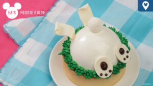 A decorated dessert shaped like a bunny, with white frosting, chocolate eyes, and green icing grass, on a pastel checkered background.