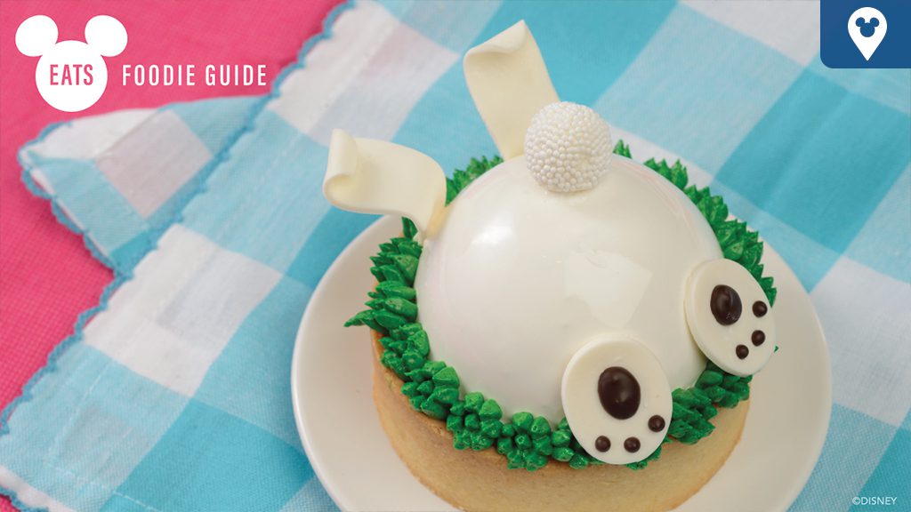 A decorated dessert shaped like a bunny, with white frosting, chocolate eyes, and green icing grass, on a pastel checkered background.