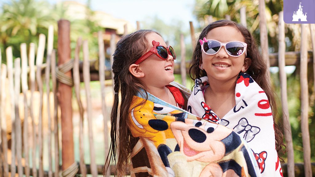 reasons-to-be-excited-disney-world Two smiling girls in sunglasses wrap themselves in blankets with cartoon characters, standing outdoors near a wooden fence.